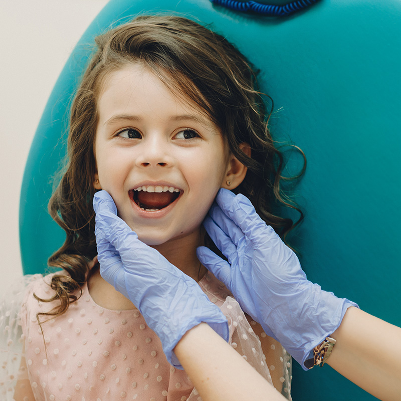 A young girl is smiling and being examined by a dental professional, with her mouth open to show her teeth.